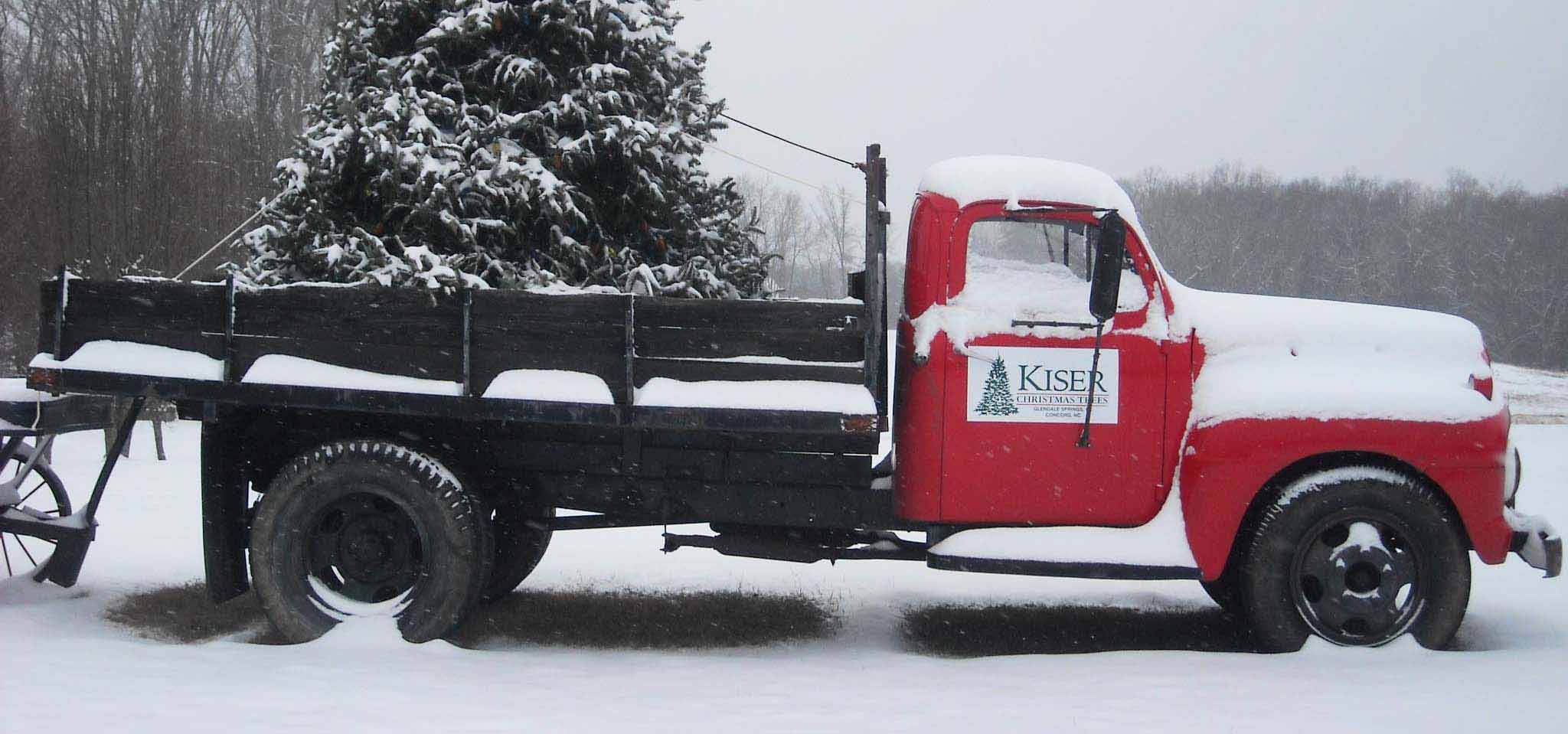 A old red pickup truck with the kiser logo carrying a fresh cut christmas tree in the snow covered mountains at christmas time.